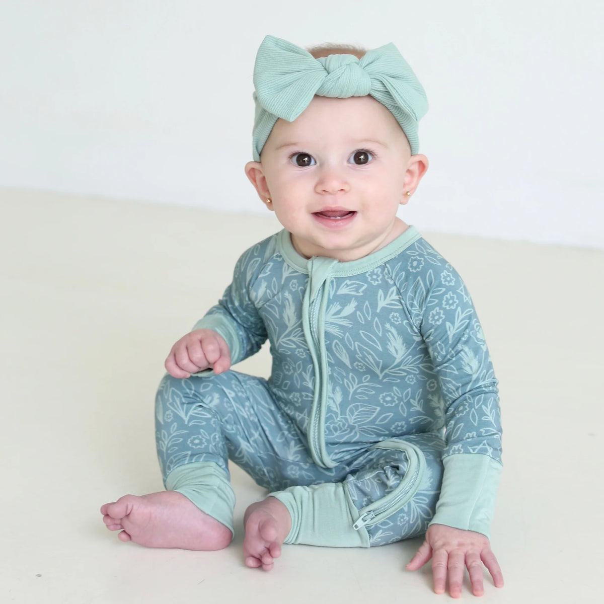 Smiling baby wearing Shiloh & Kate Sage Bloom zip-up pajamas with a delicate floral print, mint green trim, and a matching bow headband.