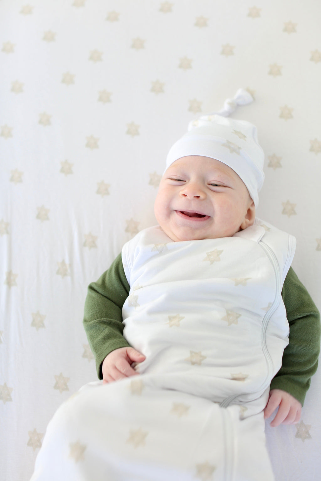 Smiling baby wearing a white Shiloh & Kate knotted gown and matching hat with a soft gold Star of David print.
